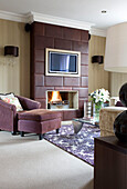 Armchair and footstool at fireside with brown leather chimney breast with plasma screen TV