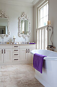 Freistehende Badewanne mit violettem Handtuch und verzierten Spiegeln über den Waschbecken in einem klassischen Badezimmer