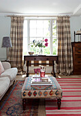Tisch am Fenster mit karierten Vorhängen im Wohnzimmer mit gemustertem Ottomanenhocker in einem Haus in East Sussex, England, UK