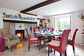 Red leather dining chairs at table with lit fire in Sussex farmhouse, UK