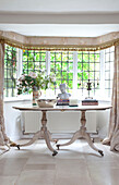 Ornaments and books on oval table in bay window of Sussex farmhouse, UK