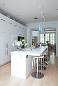 Bar stools in white fitted kitchen with blue pendant lights and view to dining room in contemporary London home, UK