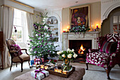 Schwarm Sessel und Weihnachtsbaum mit beleuchtetem Feuer in Surrey Bauernhaus mit geschnittenen Rosen und Teelichter auf Ottomane, England, UK