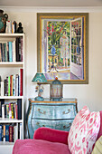 Colourful artwork above painted side unit with bookcase in living room of Sussex home England UK