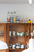 Teacups and crockery with vintage bottles in wooden glass fronted kitchen cabinet in Kent family home England UK