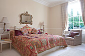 Armchair at window with colourful bed quilt and ornate mirror in West Sussex home England UK