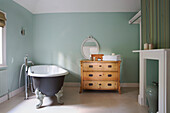 Folded towels on wooden chest of drawers with freestanding roll-top bath in light green bathroom of Berkshire home, England, UK