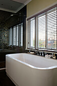 Freestanding white ceramic bath at window with venetian blinds in Berkshire home, England, UK