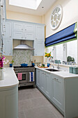 Roman blinds at window of light blue fitted galley kitchen in London townhouse, England, UK