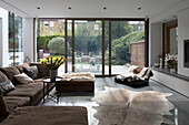 Brown sofa and footstools with animal skin rugs and view through sliding patio doors from contemporary Sussex home, England, UK
