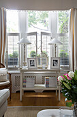 Matching lamps with family photographs on table in window with shutters,  Hertfordshire living room,  England,  UK