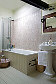 Weißes Waschbecken mit holzgerahmtem Spiegel und getäfelter Badewanne in einem gefliesten Badezimmer in Berkshire, England, UK