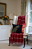Red tartan blanket on wingback armchair with black peacock cushion at window in London home,  England,  UK