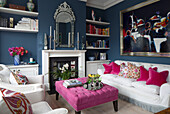 Kunstwerke und eingelassene Bücherregale mit weißem Sofa und Sesseln und rosa Ottomane in Londoner Wohnzimmer, England, UK