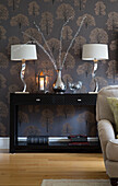 Pair of chrome base lamps on black console with patterned wallpaper in living room of Chobham home   Surrey   England   UK