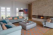 Light grey sofas with buttoned ottoman and exposed stone wall with lit fire in Lechlade home Gloucestershire England UK