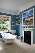 Freestanding roll-top bath at Georgian window with artwork above fireplace in Berkshire home England UK