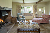 Teaset and cakes on patterned ottoman at lit fireside in Gloucestershire living room England UK