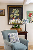 Kunstwerk und Orchideen mit hellblauem Sessel und Lampe in einem Cottage in Gloucestershire, Großbritannien