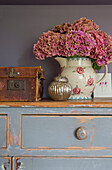 Hydrangea in floral jug with vintage glass and suitcase on chest of drawers in Alford home Surrey UK