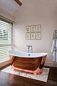 Freestanding copper bath with Venetian blinds in pastel pink bathroom with framed botanical prints in Gloucestershire barn conversion UK