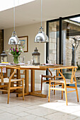Verchromte Pendelleuchten über einem Esstisch und Stühlen aus Holz in einer umgebauten Scheune in Gloucestershire UK