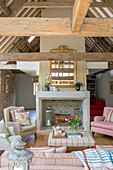 Vergoldeter Spiegel über dem Kamin mit Sesseln im Wohnzimmer mit Balken in einer umgebauten Scheune in Gloucestershire, UK