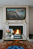 Books and hydrangeas on glass coffee table with artwork above lit fire in Wiltshire living room
