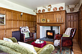 Red leather armchairs with green velvet sofa and lit woodburning stove in panelled Devon living room UK
