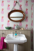 PInk pineapple wallpaper and oval mirror above pedestal basin in Hampshire home England UK