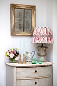 Room corner with antique painted chest and table lamp with colourful shade foxed mirror and flowers in Hampshire home UK