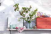 Metal bucket with branches of blossom shelf in greenhouse with book and floral print as well as cup glass vases eggs napkin and string
