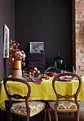 Esszimmer mit dunkelvioletten Wänden und einem herbstlich gedeckten Tisch mit goldgelber Tischdecke und Blumen und Obst der Saison