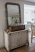 Foxed mirror with fairylights and wooden crate of bottles on painted cupboard in festive dining room