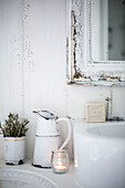 Weißes Waschbecken im skandinavischen Stil mit weißer Emaillekanne, Lavendeltopf und Teelicht, das neben einem Handwaschbecken mit Seife vor einem weißen Spiegel blinkt