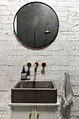 Stone washbasin with brass fittings and round mirror on a white brick wall