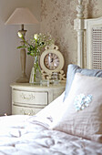 Bedside cabinet with lamp, white roses, and alarm clock with a bed in the foreground