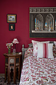 Antique bedside table next to bed with floral bedspread in bedroom with red wall