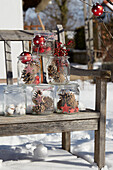 Winterliche Dekoration mit Zapfen und Beeren in Einmachgläsern auf Holzbank im Schnee