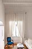 Brightly draped curtains in front of window in white room with blue chair