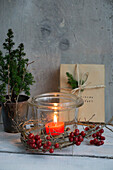 Lantern with a wreath of holly berries, behind it mini conifers and packages