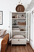 White cabinet with glass doors holding books, decorative objects, table linen, and cushions next to rattan sofa