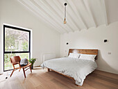 A wooden bed and a chair in a simple bedroom with a high ceiling
