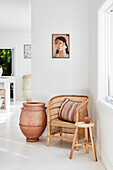 Rattan armchair, wooden stool and terracotta floor urn in corner of room, portrait painting on wall
