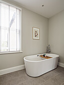 Free-standing bathtub in the bathroom with light grey walls, window with blinds