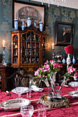 Gedeckter Tisch mit roter Damasttischdecke und Lilien, im Hintergrund antike Vitrine