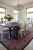 Large dining table with wooden top and white chairs on Persian carpet in bright dining area