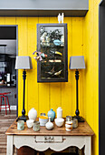 Side table with collection of vases and lamps in front of a yellow-painted wood paneled wall
