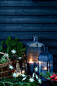 Fir branches with basket, lanterns and star-shaped fairy lights in front of wooden wall