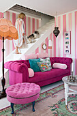 Pink sofa in lounge with pink striped wallpaper, white staircase with woman and dog